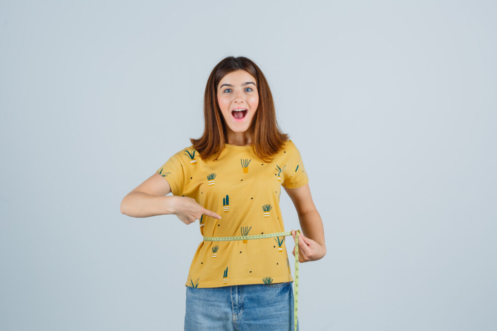 young girl measuring waist size, pointing to it in yellow t-shirt, jeans and looking jolly , front view.